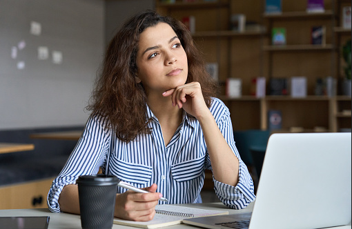 Student looking thoughtful while sitting at a desk with paper and pencil and a laptop on the desk.