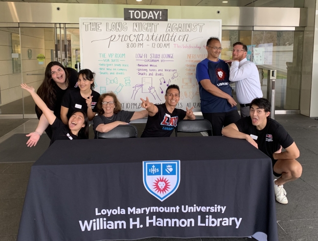 Several students and several more library staff members sitting at a table outside the library at Loyola Marymount University in California to welcome students to the Long Night Against Procrastination event. A whiteboard behind them details the event. Most people in the photo are in humorous poses or making funny faces.