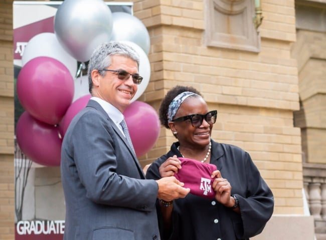 A white man and a Black woman, José Luis Bermúdez and Kathleen McElroy, together hold a Texas A&M shirt, with maroon and silver balloons behind them.