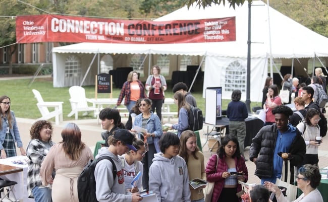 Students gather in small groups at at tables on the grounds of Ohio Wesleyan University.  A banner saying "Connection Conference" hangs in the background.  