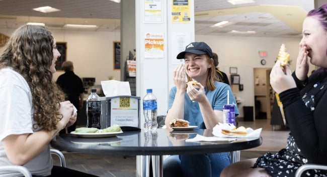 Three female students sit at a small table in a cafeteria eating sandwiches, laughing.