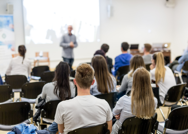 Students sit in a lecture hall facing the instructor at the front of the room