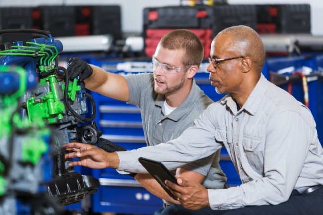 A man shows another man in goggles how to operate a machine. 