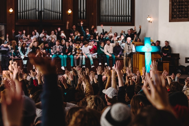 A crowd of students and visitors hold their hands up at Asbury University's chapel as someone preaches from a podium with a large blue cross.
