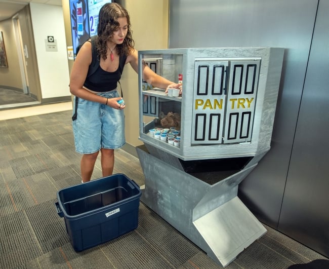 A student wearing a backpack helps restock a gray food pantry from a blue plastic tub.