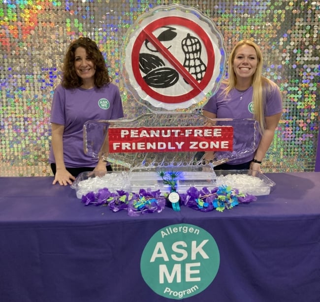 Two Duke Dining staff members smile at a table featuring a peanut-free friendly zone ice sculpture to share the launch of the Ask Me program.