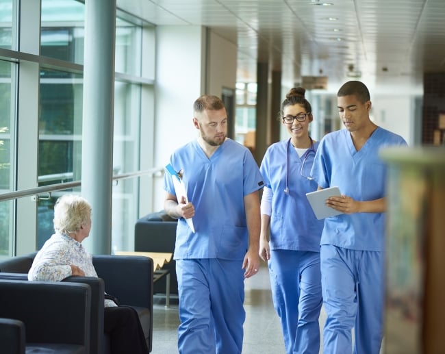 Three students in blue scrubs walk down a hallway in a hospital