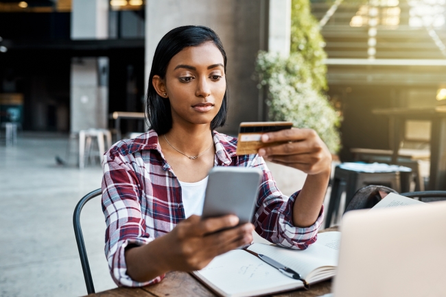 A young woman in a red flannel holds up her credit card as she makes a purchase on her cellphone.