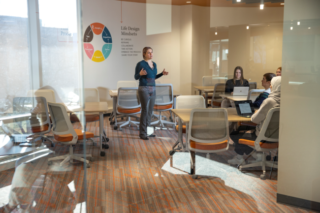 A woman stands in a classroom talking to students on their laptops in front of a wall feature a circle with the six Life Design principles.