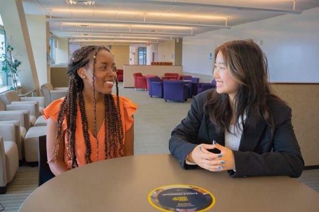 Two UCSD students smile at one another, sitting at a round table in an academic building