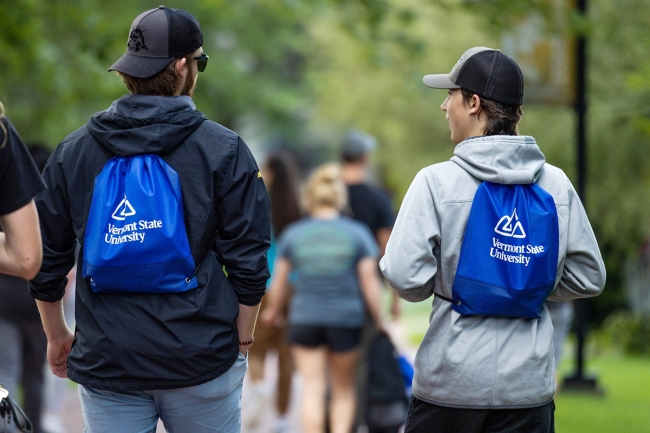 Two students in baseball caps and hoodies walk side by side down an outdoor path wearing Vermont State U–branded drawstring bags.