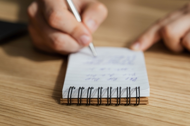 A man's hands are showing taking notes in a small spiral notebook on a wooden desk.