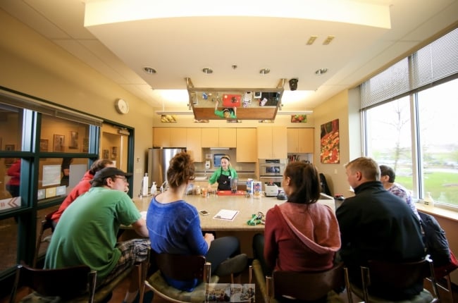 Wide-angle photo of students gathered around a large table at the University of North Dakota's test kitchen. A young female chef/instructor is demonstrating something with a blender toward the back of the table. Ceiling holds a mirror for better views of the demonstration.