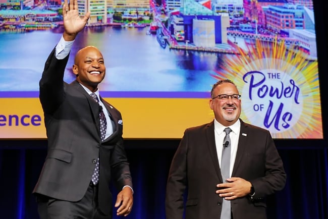 Two men in suits, one younger, Black, and bald, the other a little older, Hispanic, and wearing glasses with a short white goatee, waving on stage