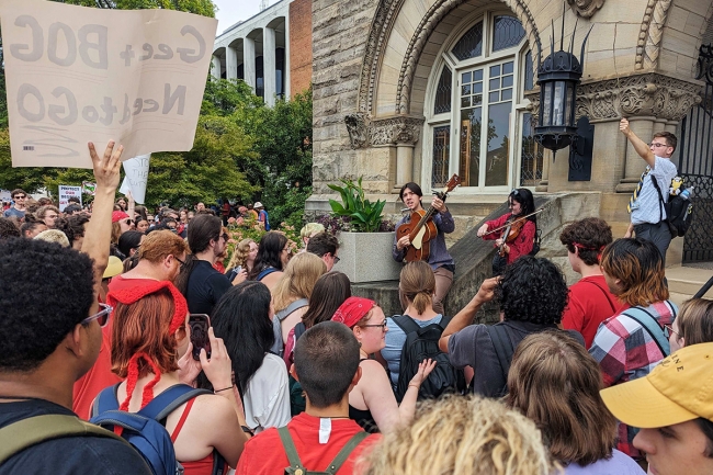 West Virginia University students protesting proposed faculty member layoffs and academic program cuts. One student holds a stick above the crowd, and another has a sign reading "Gee + BOG Need to GO."