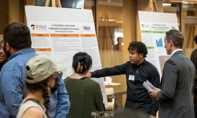 Two students in a large room share their science research poster with a professor as others walk by.