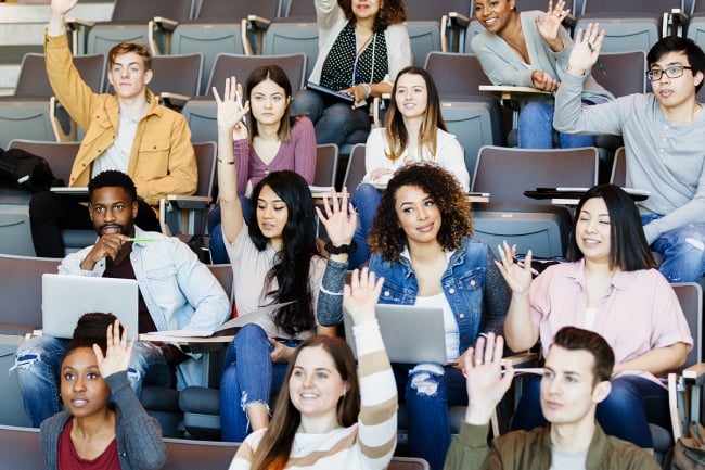A group of students in a lecture hall raises their hands. 