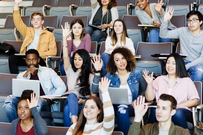 A group of students in a lecture hall raises their hands.