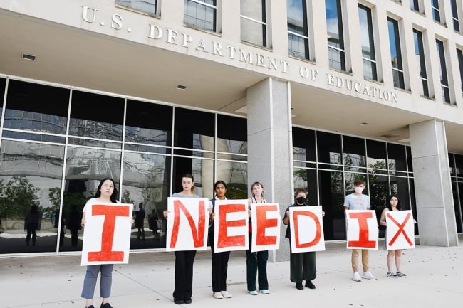 Seven students hold white poster boards painted with red letters that spell I Need IX.