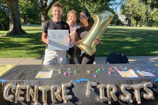 Three students stand at a black table with golden balloon letters that read "Centre Firsts"