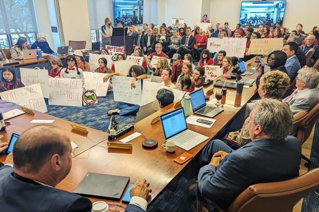 Over 100 people, including students covering the floor and holding signs, fill the West Virginia University Board of Governors meeting room.