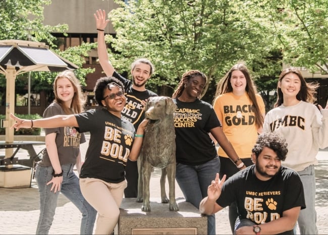 A group of happy-looking students wearing black and yellow-gold swag and gear pose on the University of Maryland Baltimore County campus, presumably for orientation.