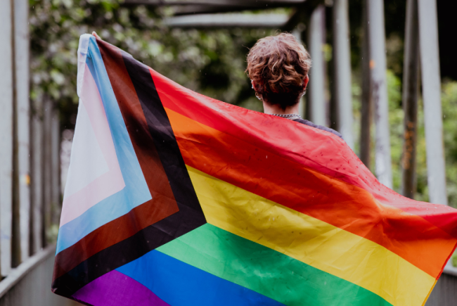 A young person carries a Progress Pride flag unfurled behind their back. 