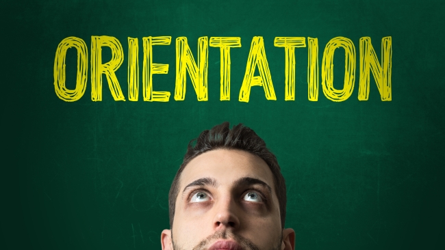 A male student looks up at the word "orientation" written in yellow on a green chalkboard behind him.