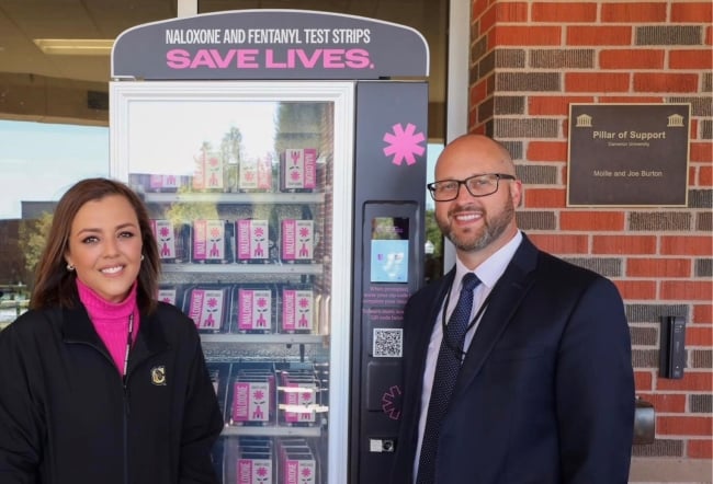 Two individuals stand next to a pink-and-black vending machine containing Narcan.