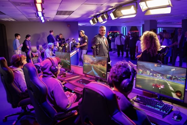 Gamers with headsets sit at a row of computers while others stand in the background of the new esports arena at Brookdale Community College. Lighting is dim and purplish-red.