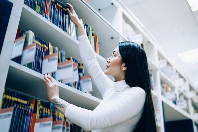 Asian woman picks a book off of a shelf at a library