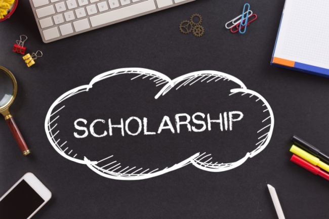 A graphic with the word "scholarship" in a cloud-shaped bubble, against the background of a desk, with a keyboard and school supplies visible in the background. 