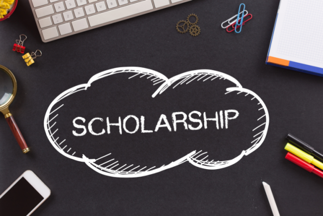 A graphic with the word "scholarship" in a cloud-shaped bubble, against the background of a desk, with a keyboard and school supplies visible in the background.