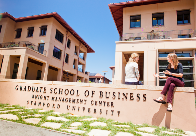 Two students sit on a Stanford Graduate School of Business sign on the Palo Alto campus.