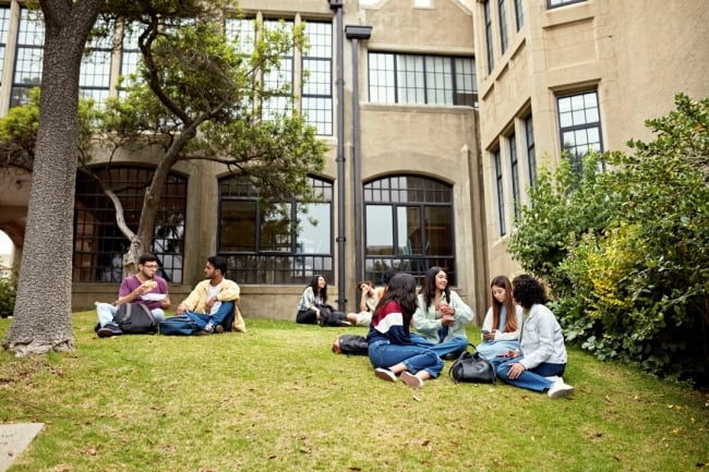 Students sit on a lawn outside of a college building.