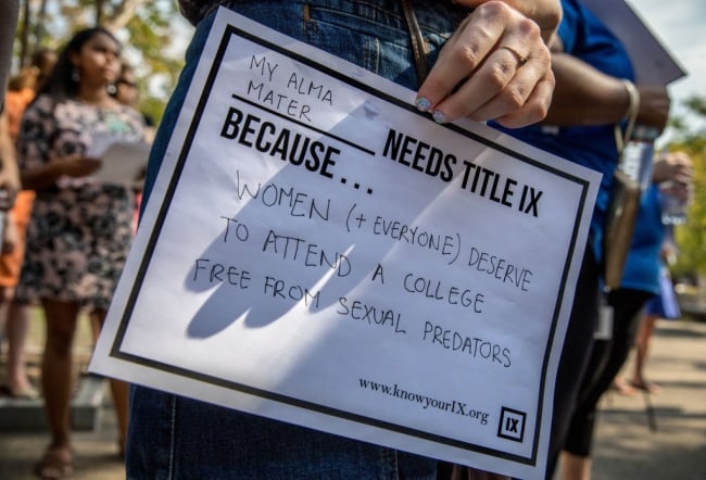 A person holds a sign on white paper that says "My alma mater needs Title IX because women and everyone deserve to attend a college free from sexual predators."