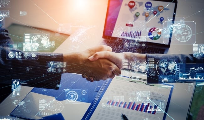 Two male hands shake while there are blue "digital" icons floating around. A computer screen and paper bar chart are in the background.