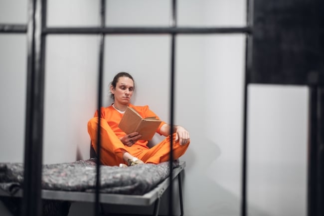 A man in an orange jumpsuit sits on a bed reading a book behind bars. 