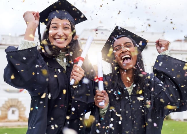 Two female, smiling students celebrating in caps and gowns outside on graduation day. They're throwing confetti, with diplomas in hand.