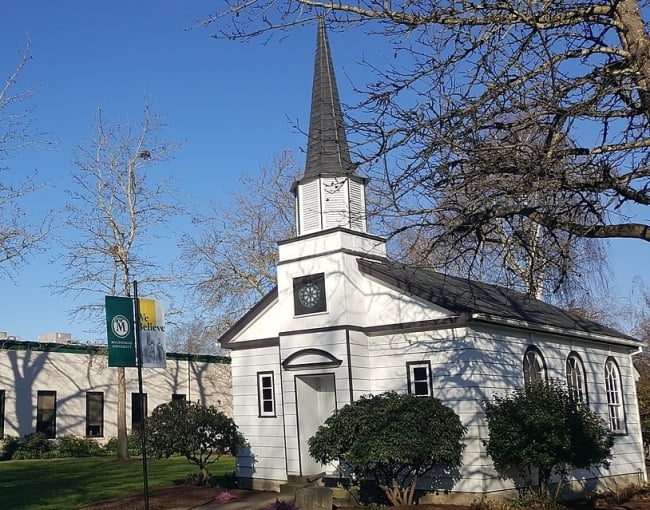 An exterior view of the Multnomah University campus showing trees, blue sky and a white building with a steeple.
