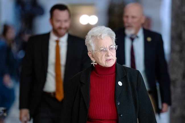 Virginia Foxx, a light-skinned woman with white hair wearing a red top under a black blazer, walks in a hallway.