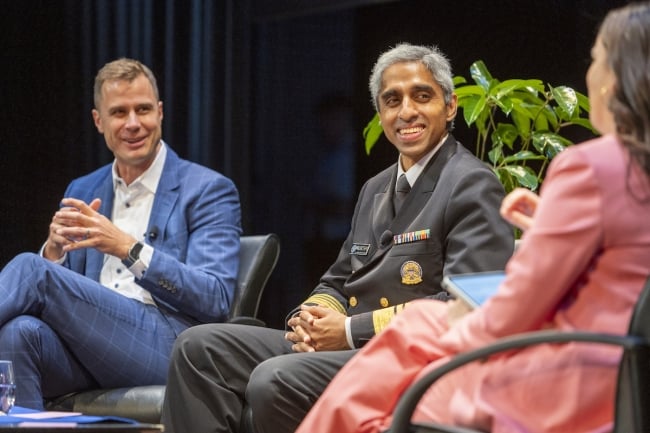 Surgeon General Vivek Murthy, a brown-skinned man with bright gray hair wearing the traditional military uniform of the surgeon general, is flanked by Kate Bowler, a professor of American religion at Duke, who is wearing pink, and men’s basketball coach Jon Scheyer, a light-skinned man wearing a blue suit, onstage at the Page Auditorium at Duke University.