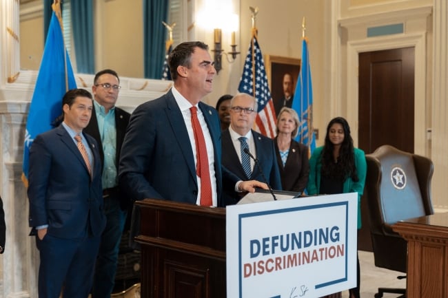 A man in a suit stands at a podium flanked by supporters and an American flag. A sign on the front of the podium says "Defunding discrimination."
