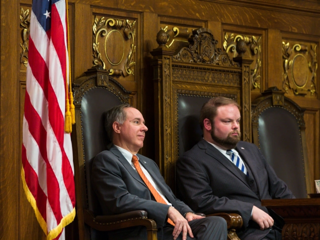 Two men sit in a state legislative chamber, flanked by American flags