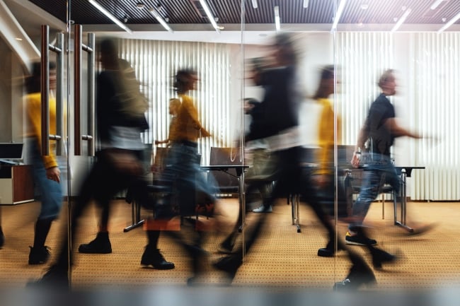 People walk quickly (their faces and bodies blurred due to motion) inside an office. 