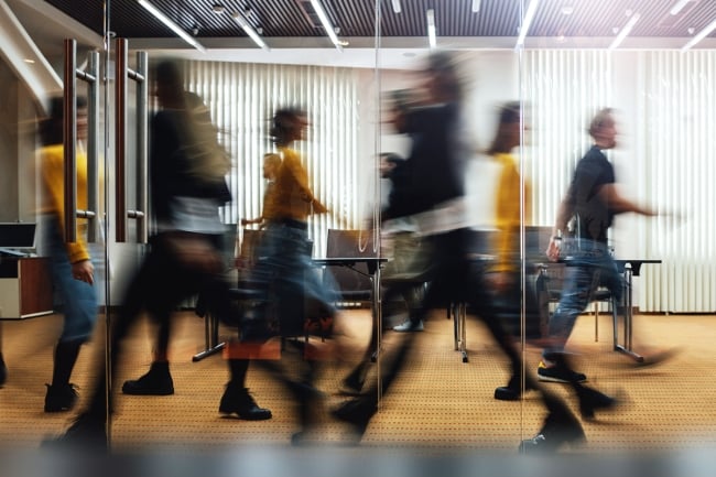 People walk quickly (their faces and bodies blurred due to motion) inside an office.