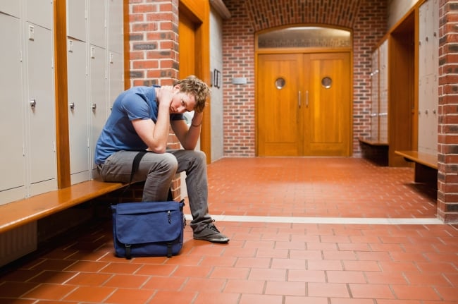 A male college student sits alone in a hallway with his hands on his head, looking anxious and stressed.