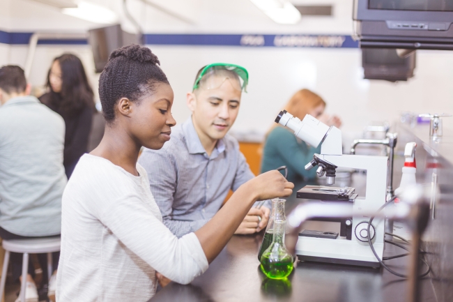 Two college students work together in the chemistry lab. She is carefully measuring solutions into two different beakers. He is watching closely. Other students are working in the background.