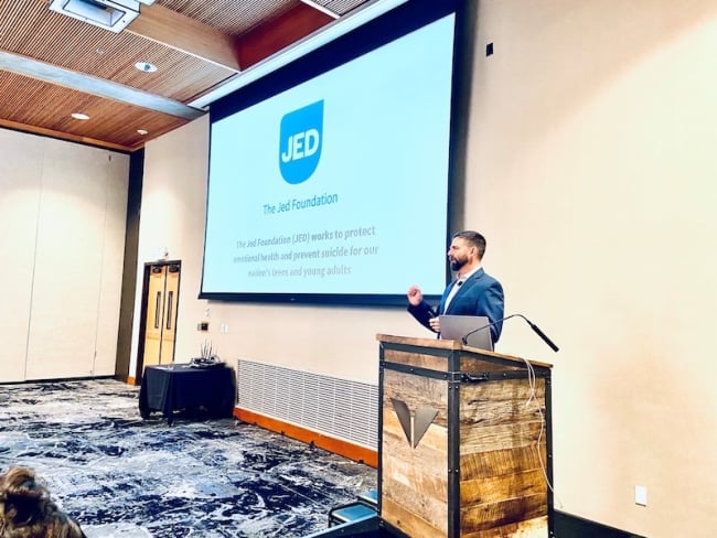 A man with a beard stands at a podium in front of a PowerPoint with the JED logo on it.