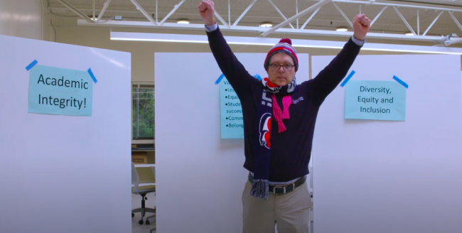 A man stands in front of whiteboards with signs taped to them reading "academic integrity" and "diversity, equity and inclusion" with both hands raised excitedly.